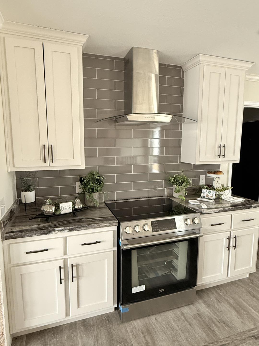 Modern kitchen with white cabinets, stainless steel oven, and hood. Gray subway tile backsplash and dark countertops. Potted plants add a fresh touch.