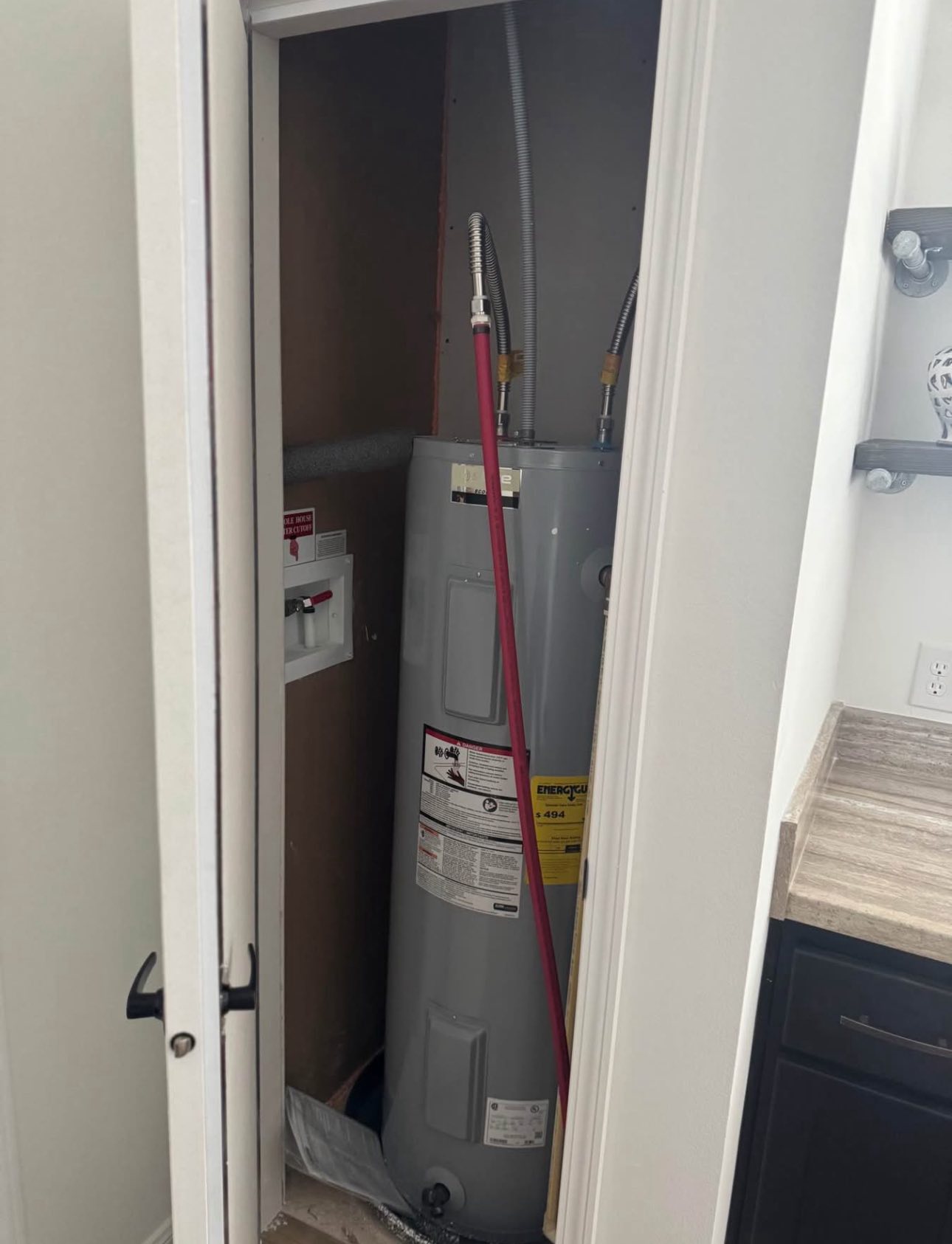 Alt text: "A water heater installed in a narrow utility closet. The closet door is partially open, revealing pipes and a control panel. Nearby, a countertop and cabinet are visible, suggesting a laundry or utility room setting. The scene conveys a functional, orderly space."