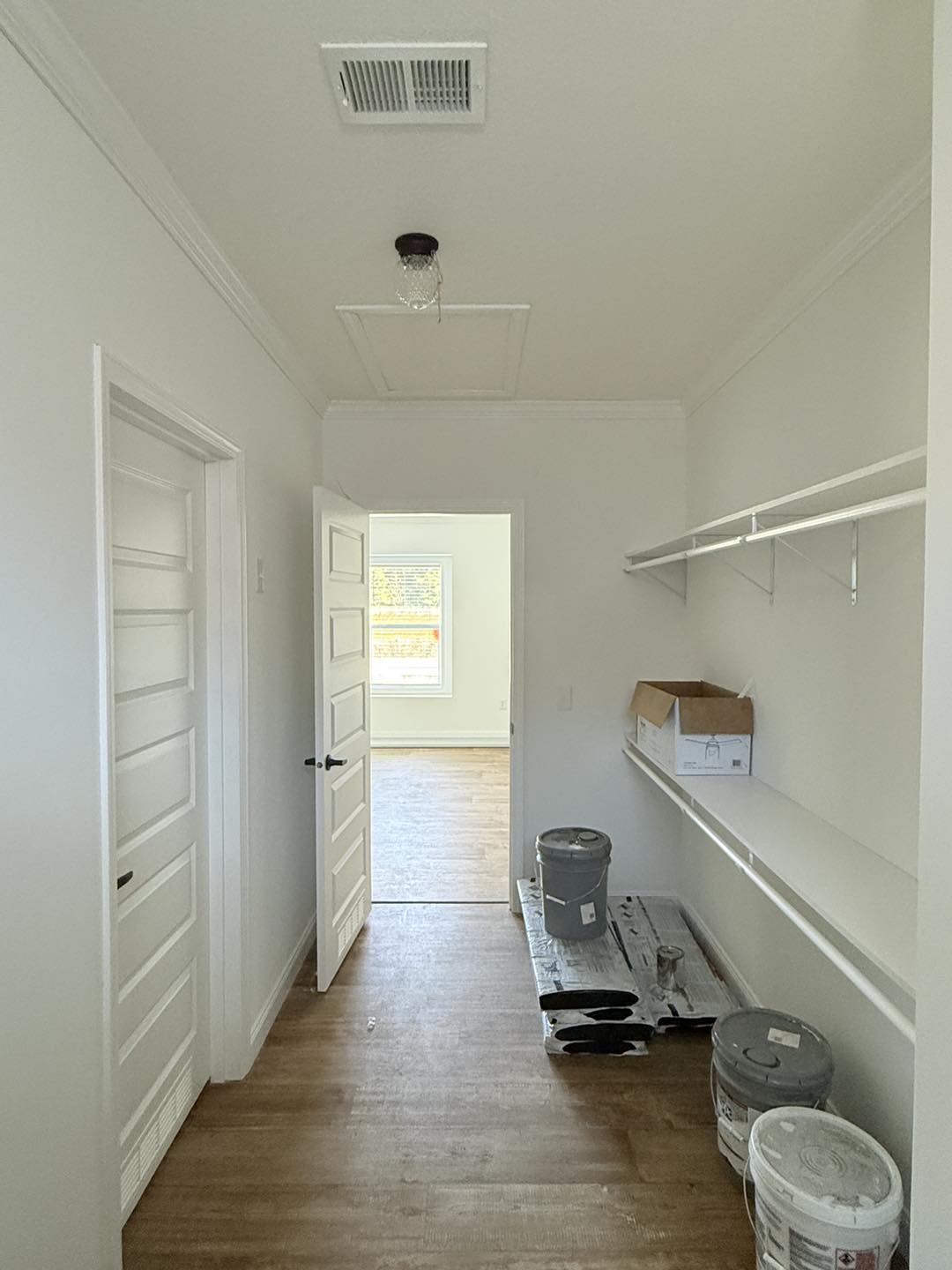 A narrow, freshly painted hallway with a wooden floor leads to an adjoining bright room. Shelves and paint cans suggest ongoing renovation, conveying a fresh start.