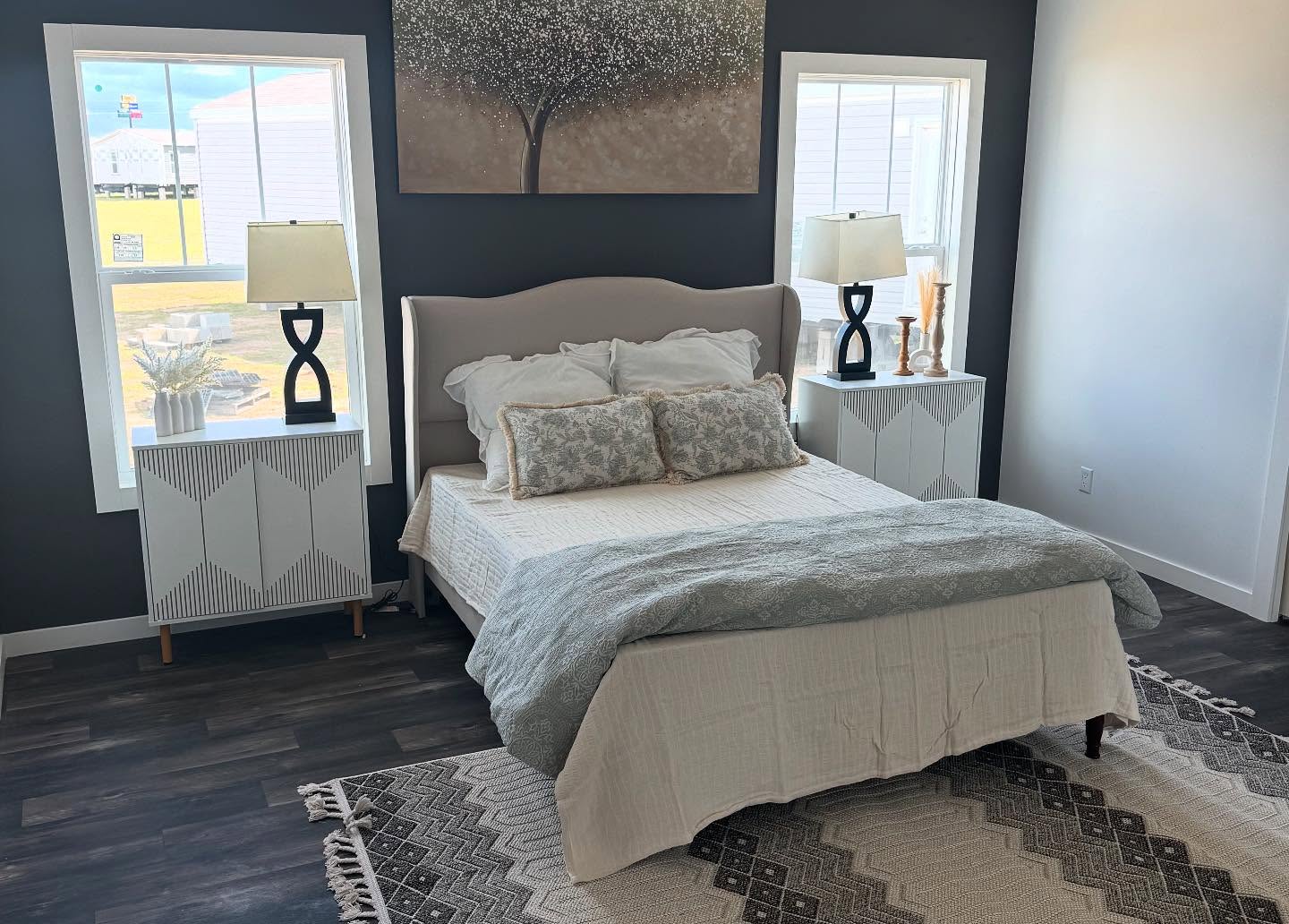 A cozy bedroom with dark walls features a double bed with a gray headboard, patterned pillows, and a textured blanket. Two windows frame the bed, illuminating abstract tree artwork above. White geometric nightstands with modern lamps and decor complete the serene setting.