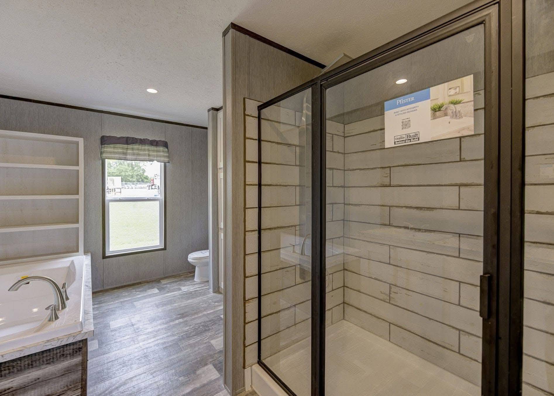 Modern bathroom with a glass shower, white-tiled walls, and wood flooring. A bathtub and shelves are on the left, with a window and toilet in the background.
