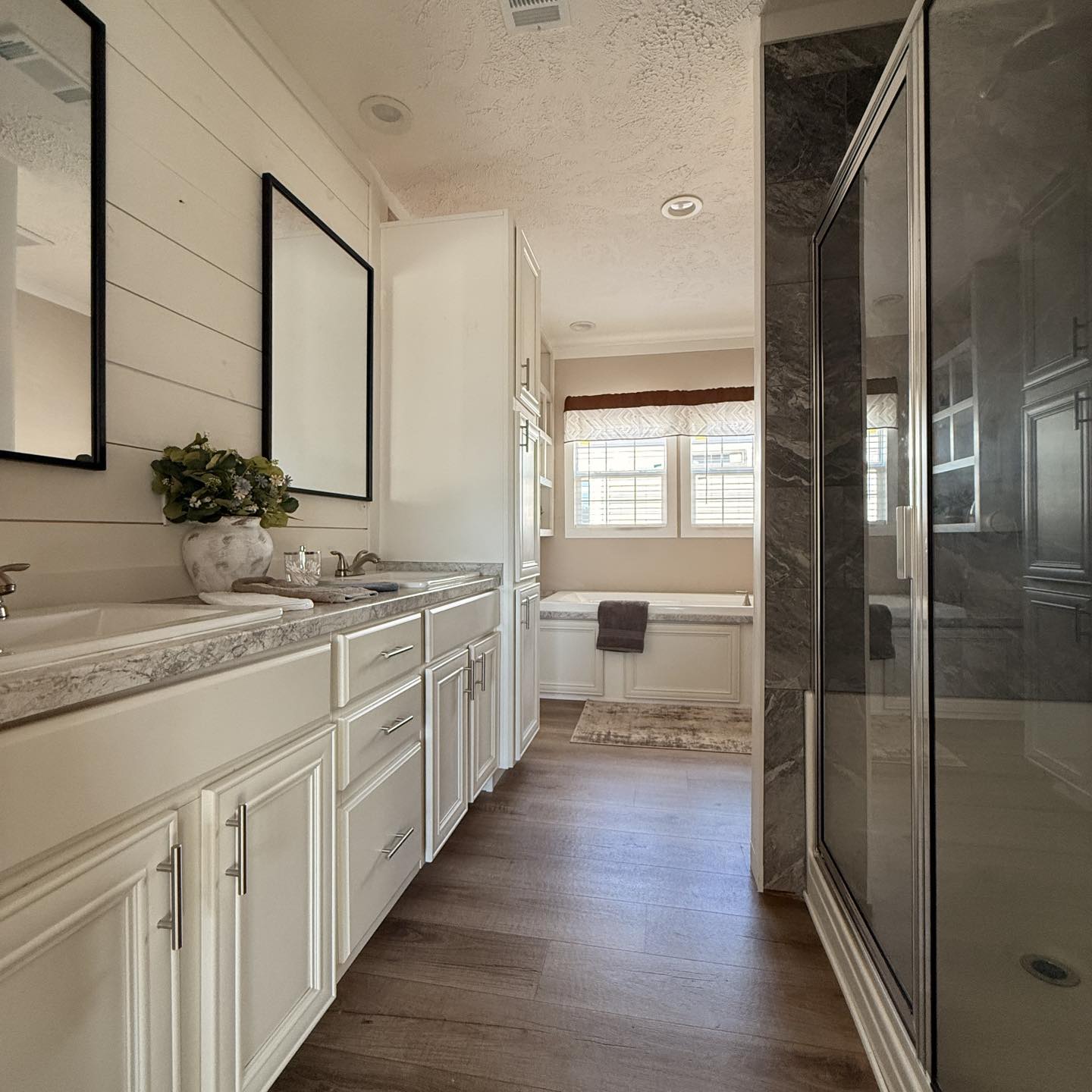 Modern bathroom with dual sinks, marble countertops, and two large mirrors. A glass shower enclosure and a sunlit bathtub area with a cozy bench are visible. The space is bright and elegant, with white cabinets and wooden floors.