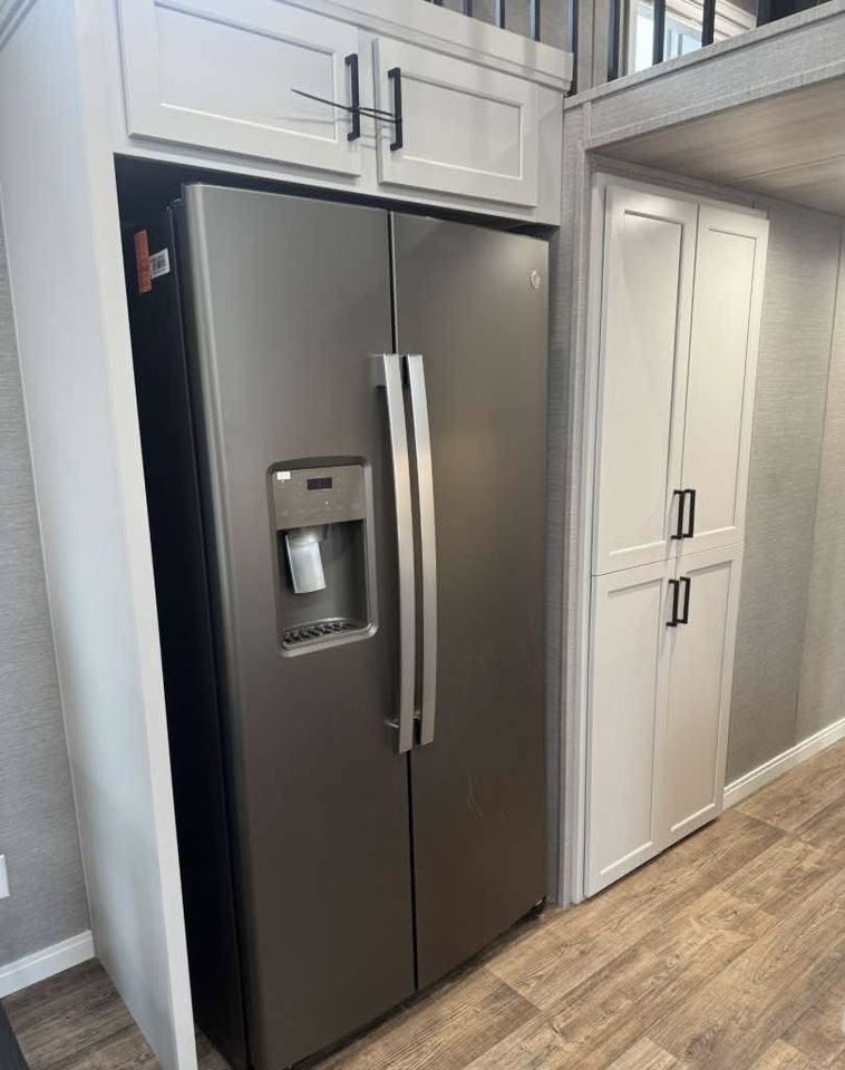 A modern, stainless steel double-door refrigerator with a water dispenser is set between white cabinets and light wood flooring in a bright kitchen.