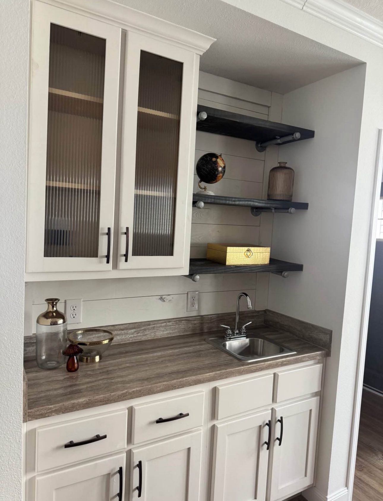 A modern kitchenette features white cabinets with vertical glass panels, a gray countertop with a small sink, and dark floating shelves with decor.
