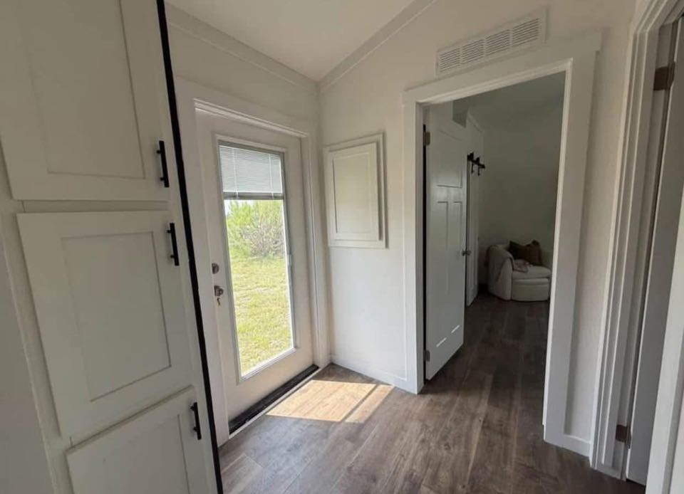 Bright hallway with white walls and wood flooring leading to a cozy room with a white armchair. Sunlight streams through a glass door, creating a calm ambiance.