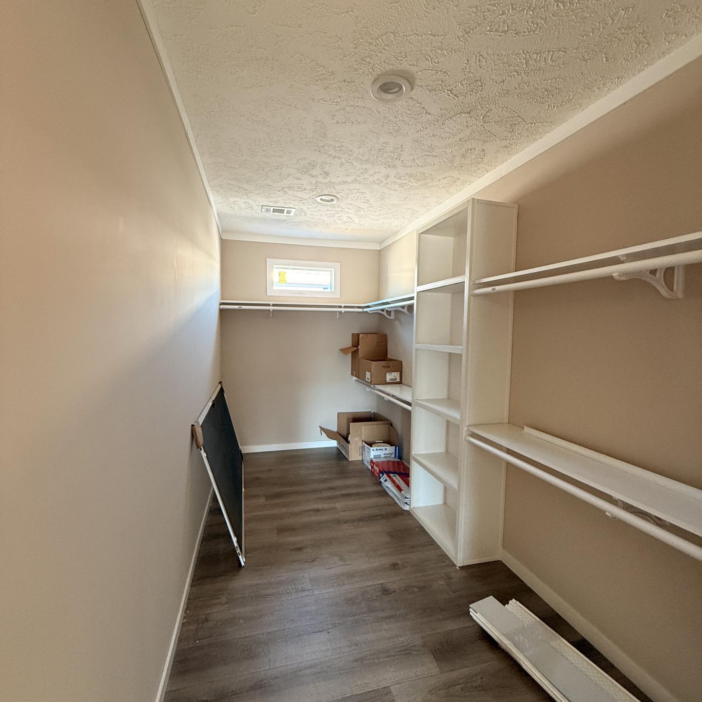 A narrow, empty walk-in closet with white shelves and rods, beige walls, and a wooden floor. Boxes are stacked neatly on the shelf, under soft natural light.