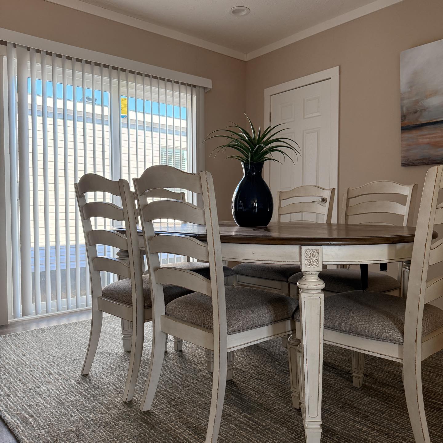 A cozy dining room with a round wooden table and six beige chairs. A dark vase with green leaves is centered on the table. Sunlight filters through vertical blinds, creating a warm and inviting atmosphere.