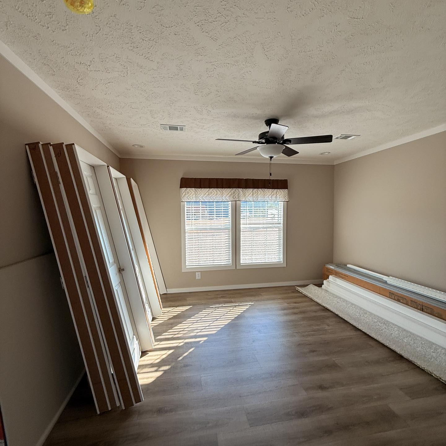 Empty room with beige walls and wooden floors, featuring a ceiling fan, windows with blinds and valance, and stacked doors and materials on the side.