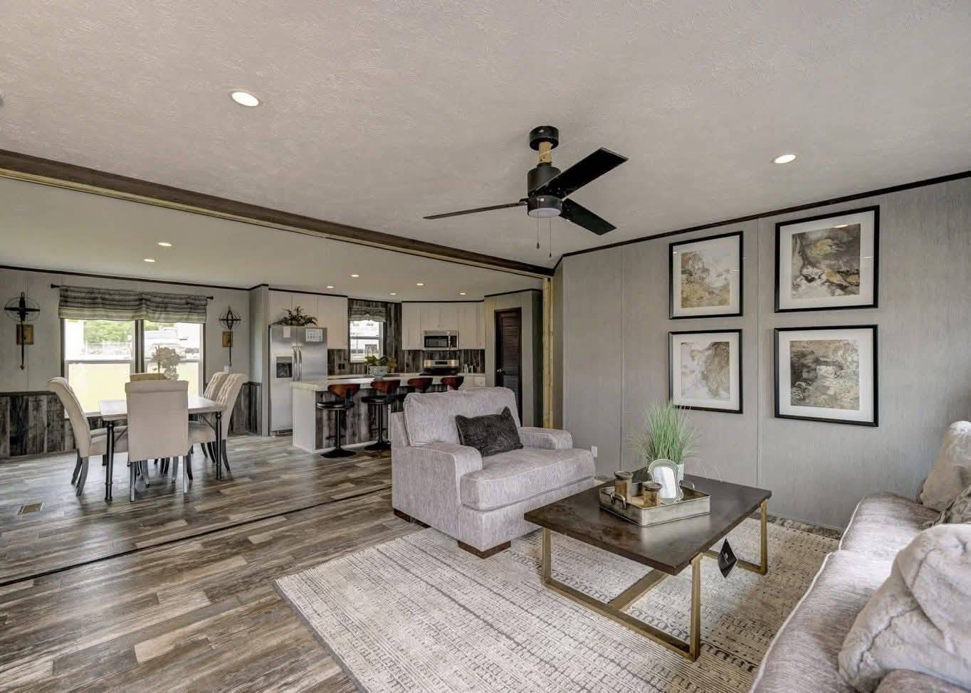 Modern open-concept living room and kitchen with neutral tones. The room features cozy gray furniture, framed wall art, a ceiling fan, and a dining area with a window view.