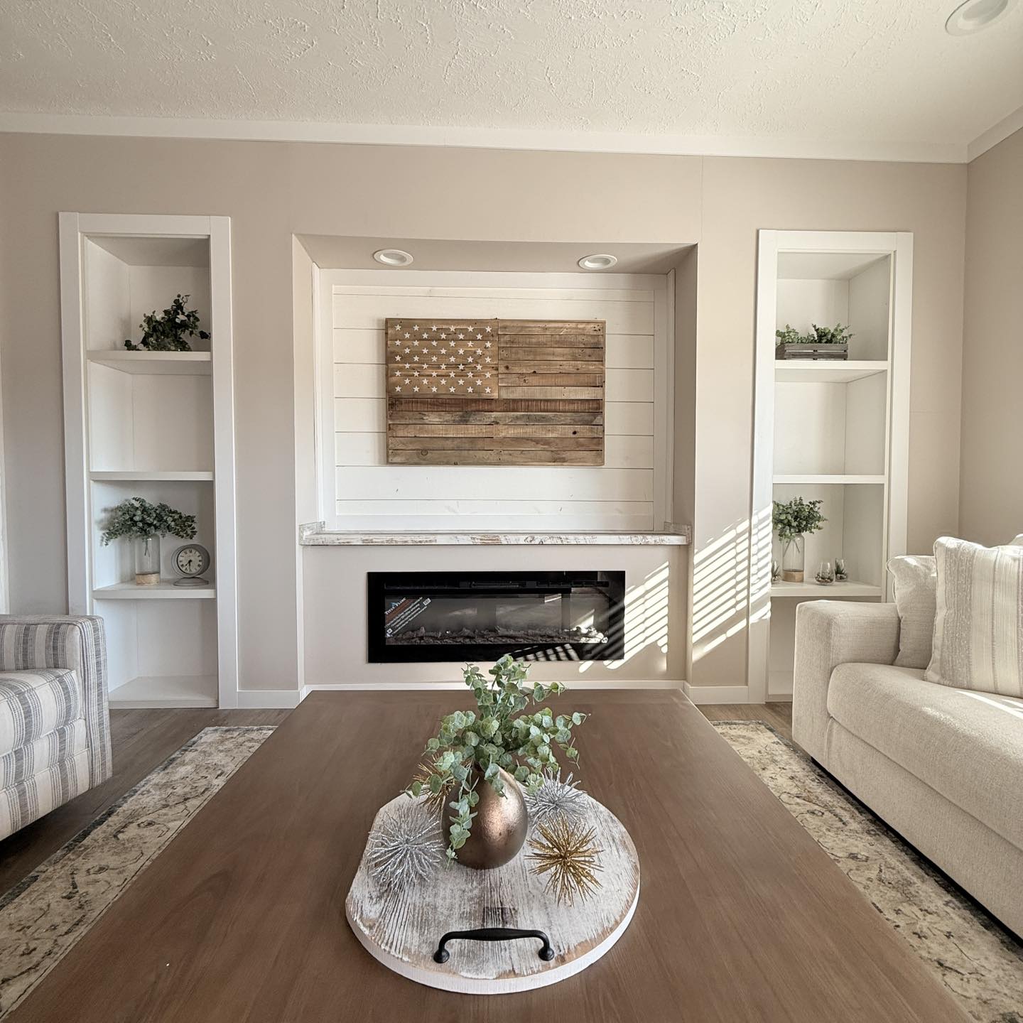 Modern living room with neutral tones, featuring a striped couch and beige sofa. A wooden flag hangs above the fireplace, flanked by shelves with plants. Cozy ambiance.
