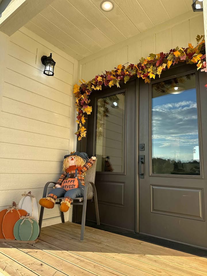 Front porch with fall decor, featuring a plush scarecrow in a chair, fall leaf garland around the door, and decorative pumpkins, under soft lighting.