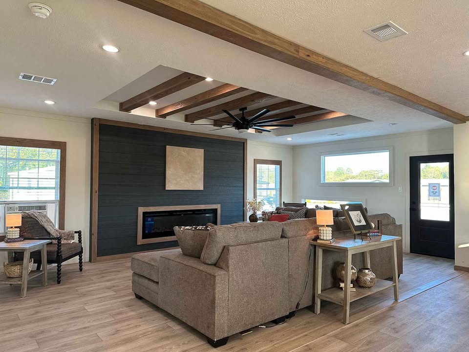 Modern living room with a cozy ambiance. Features gray sofas, wood ceiling beams, a dark accent wall with a fireplace, and large windows for natural light.