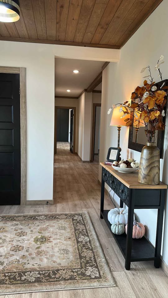 A cozy hallway with a wooden ceiling and light wood flooring features a black console table adorned with a lamp, autumn leaves, and framed items. Pumpkins beneath the table add a seasonal touch. A beige patterned rug lies on the floor, enhancing the warm and inviting ambiance.