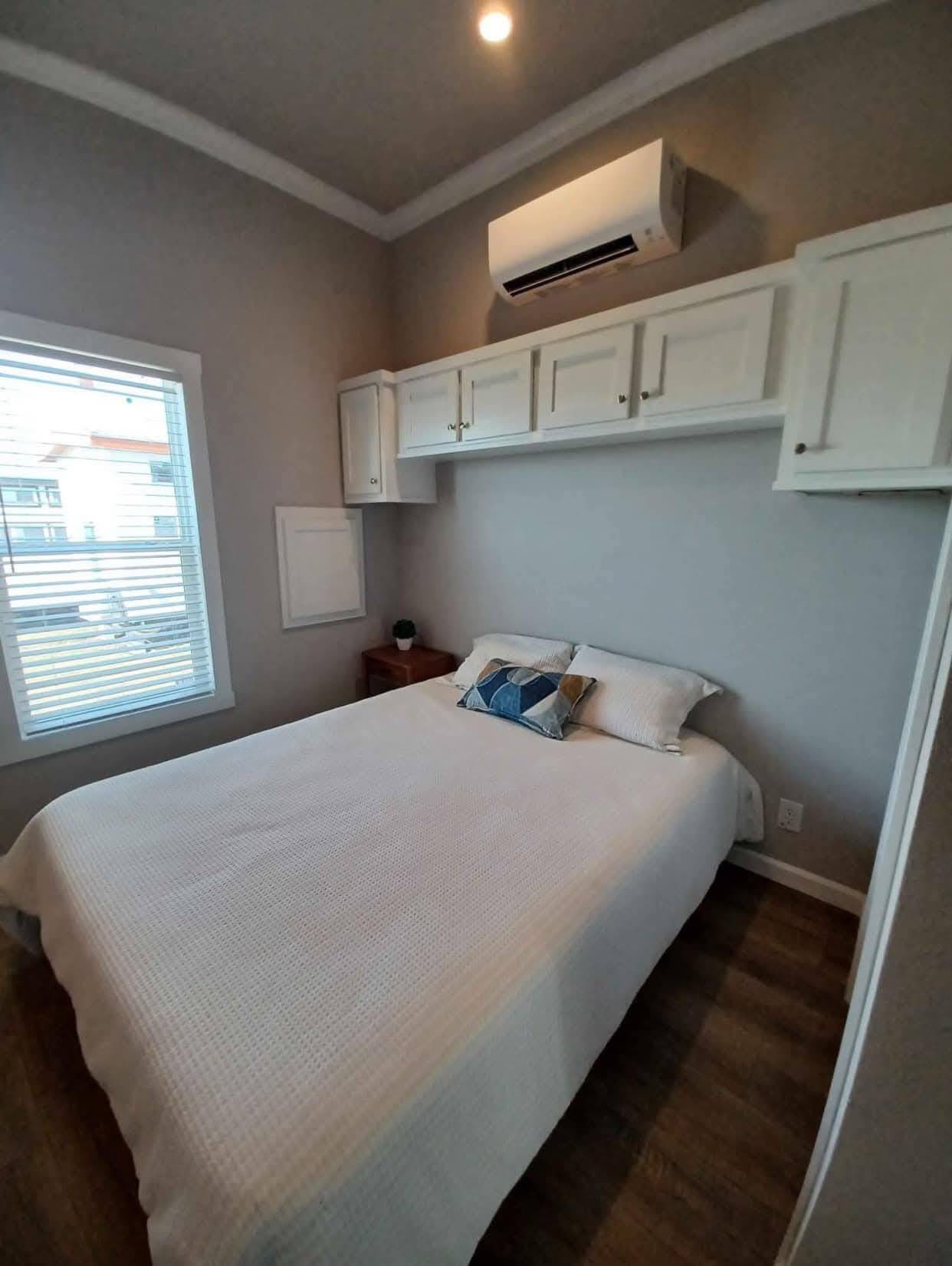 Small, cozy bedroom with a neatly made bed, white comforter, and decorative pillows. White cabinets and an air conditioner hang above. Minimalist decor.
