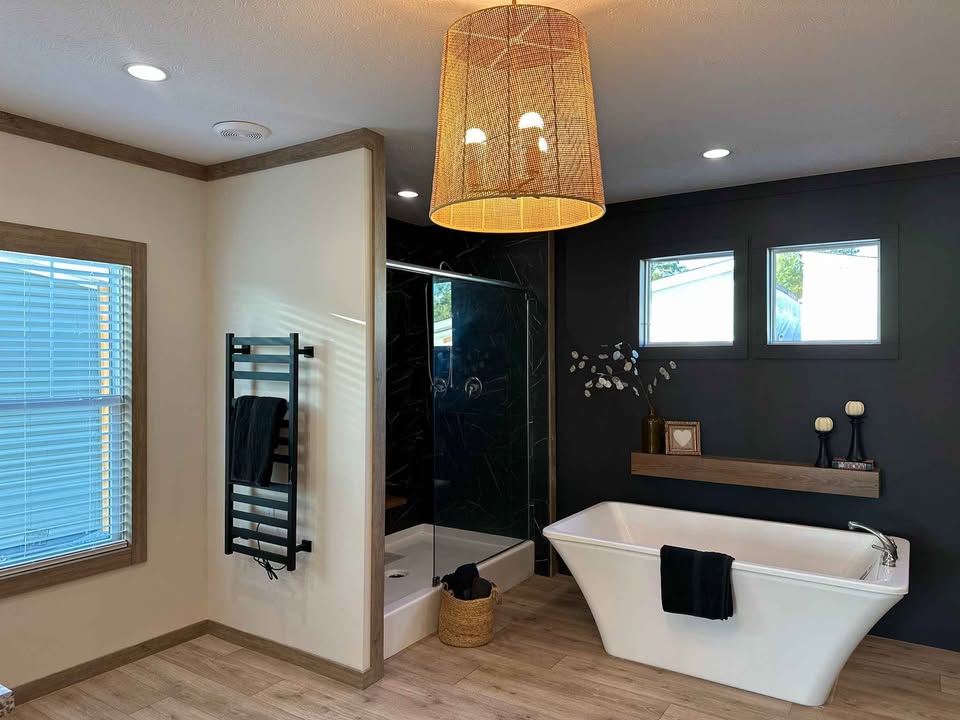 Modern bathroom with a freestanding bathtub, black accent wall, and glass shower. A large wicker light fixture hangs above warm wooden floors.