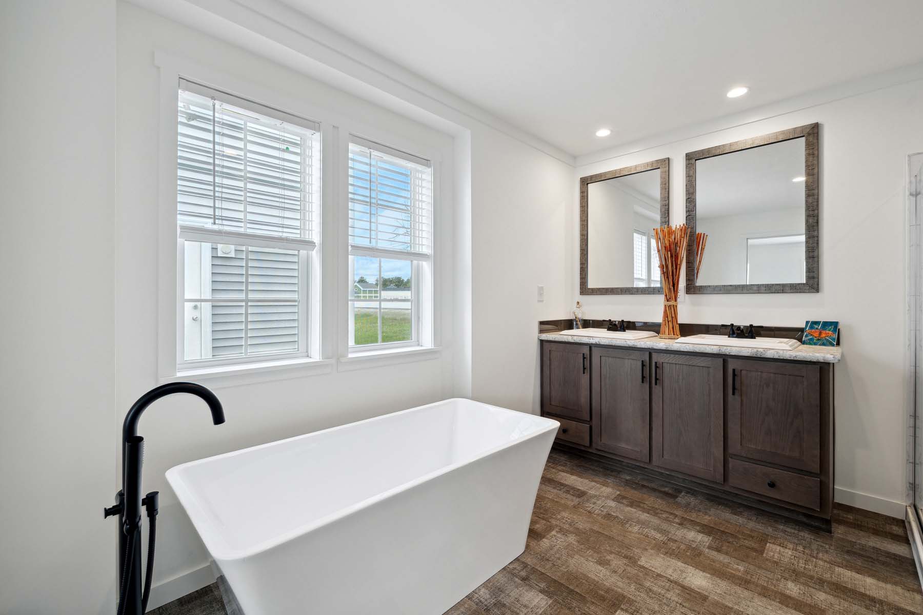 Modern bathroom with a white freestanding bathtub, sleek black faucet, and large window. Double vanity with dark wood cabinets, twin mirrors, and warm lighting.