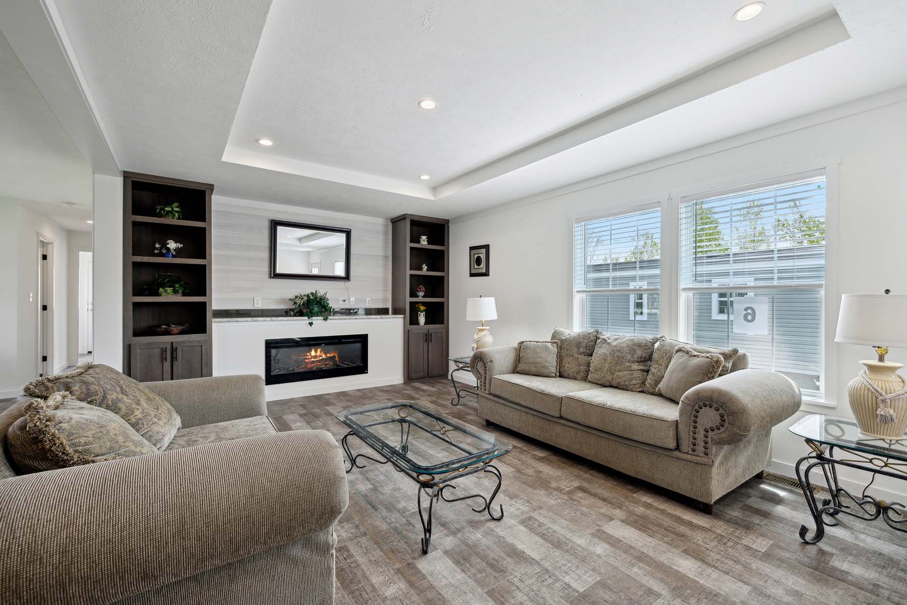 Modern living room with beige sofas, glass coffee table, and built-in shelving around a recessed fireplace. Large windows allow natural light in.