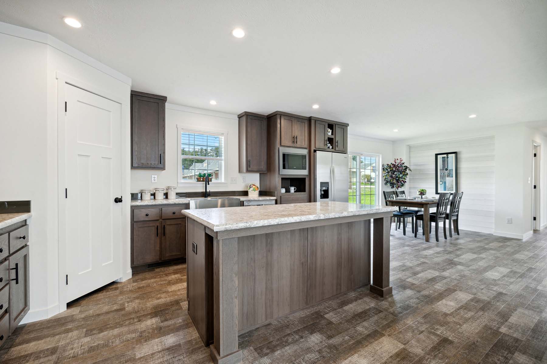 Modern kitchen interior with wooden cabinets, a large island with marble countertop, and stainless steel appliances. Dining table visible in the background.
