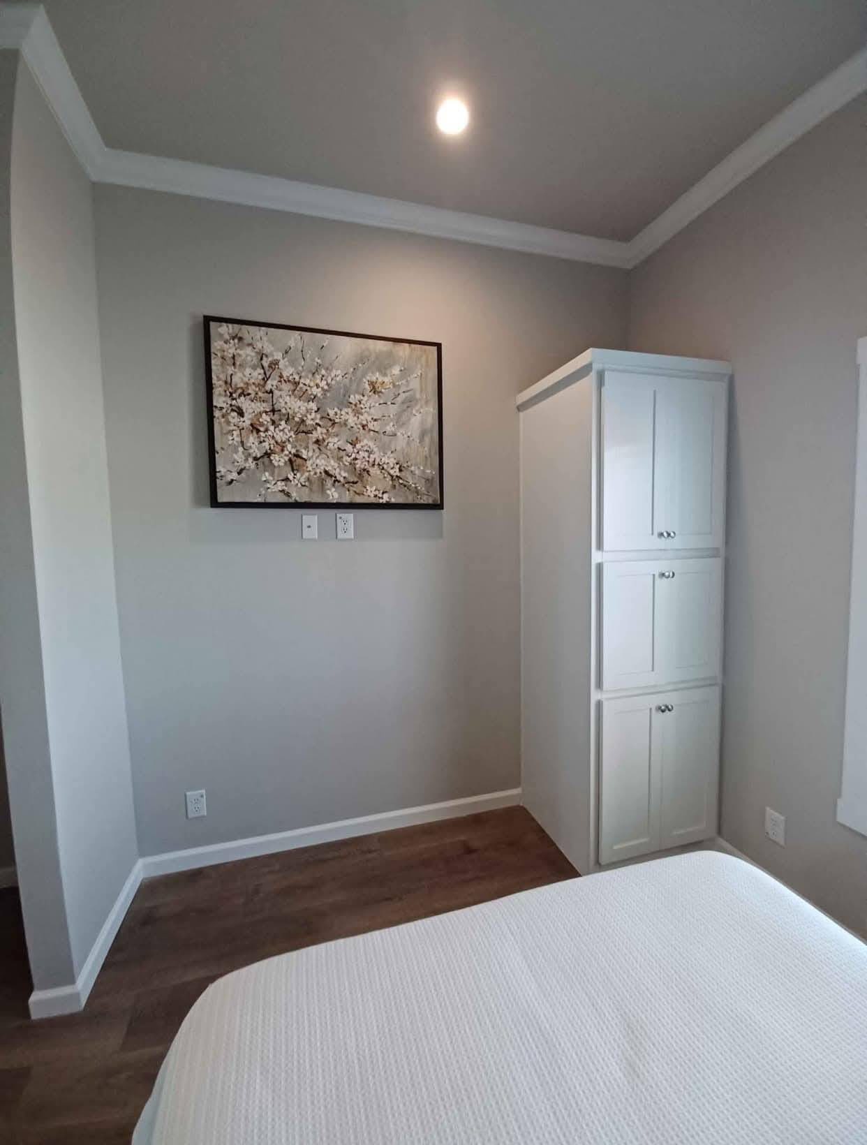 Minimalist bedroom with light gray walls, a floral painting, soft lighting, and wooden floor. White dresser and bed with a textured white cover. Calm ambiance.