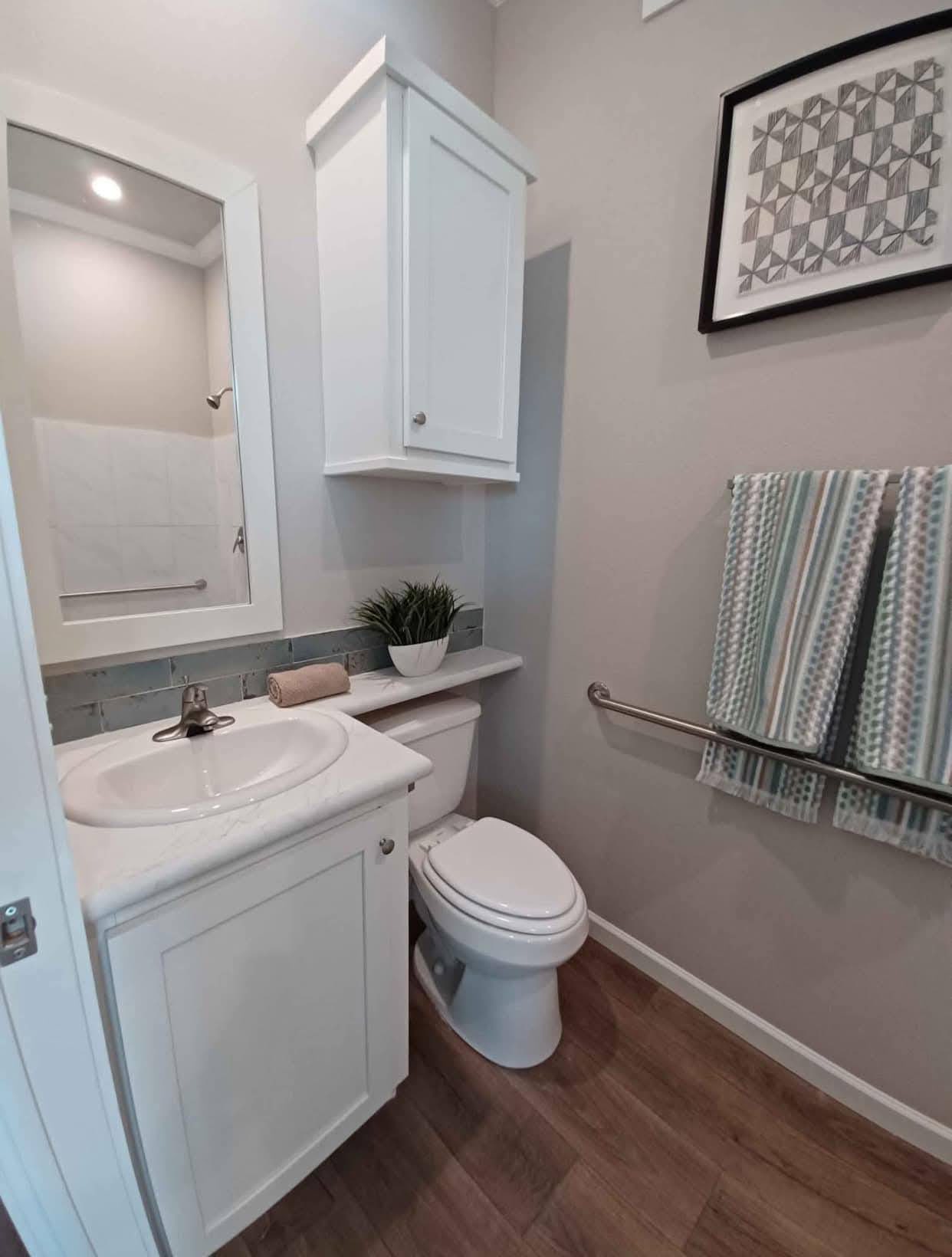 Bathroom with a white vanity, mirror, and upper cabinet. A toilet is beside a shelf with a plant. Striped towels are on a rail, and the decor feels modern.