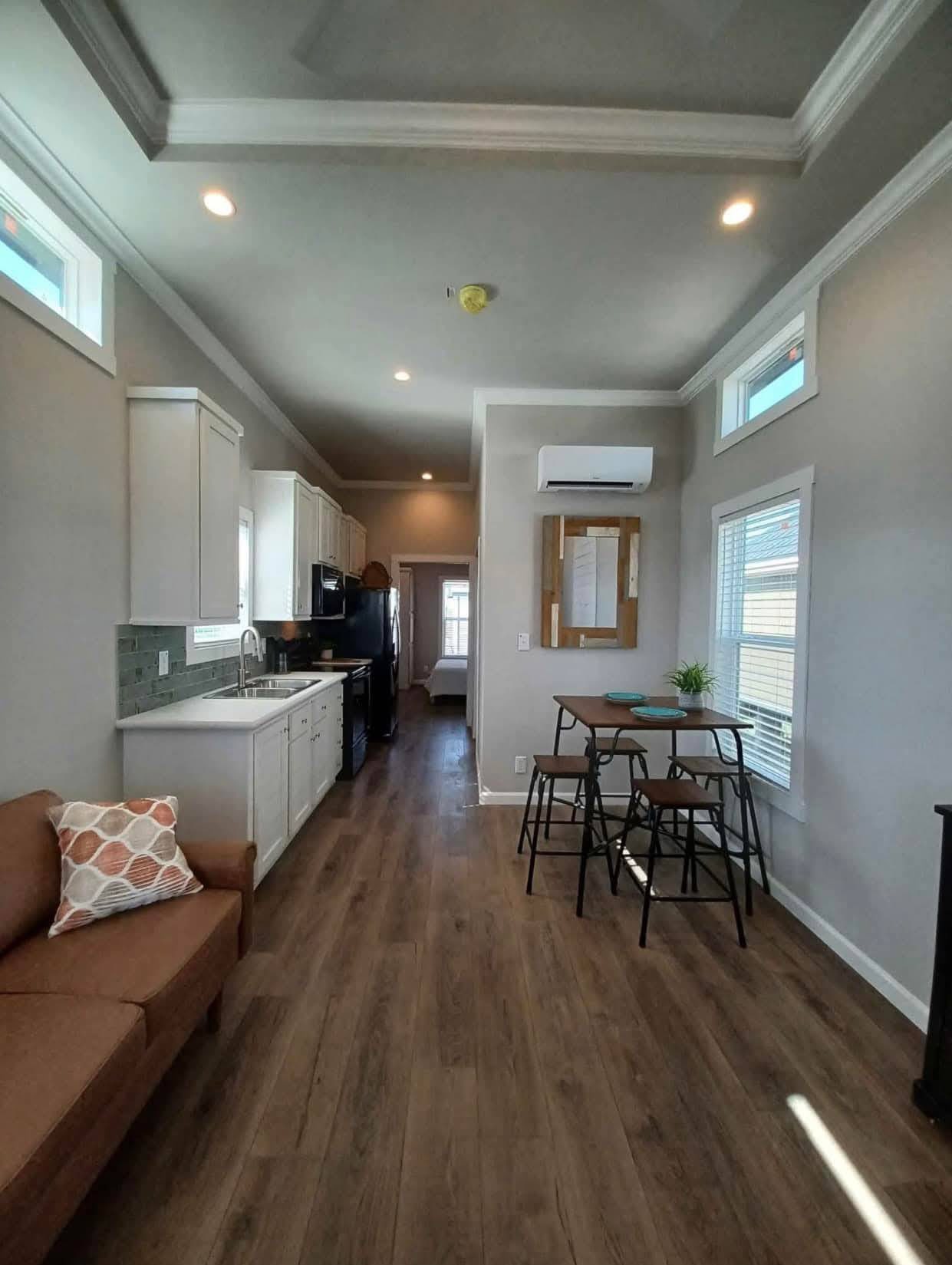 Modern, narrow living space with a brown sofa, small dining table, white kitchen cabinets, and wood flooring. Bright and cozy atmosphere.