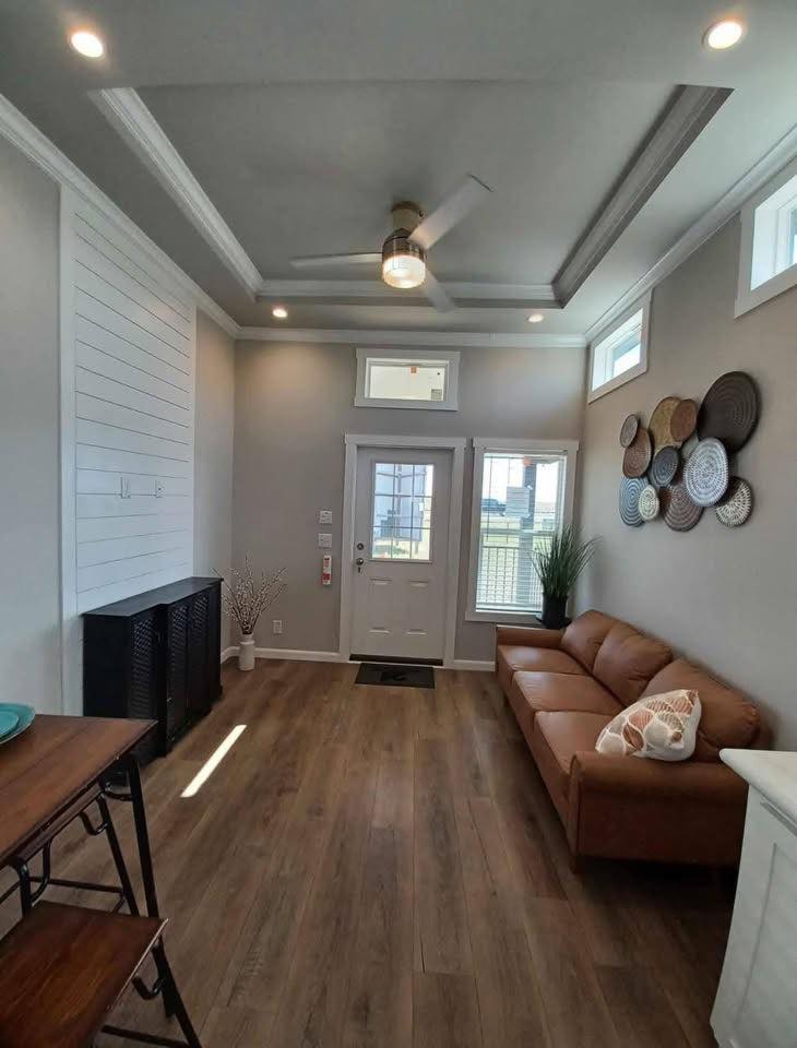 Cozy living room with high ceiling and recessed lighting. Features a brown leather sofa, decorative wall baskets, and a door with sidelights. Warm, inviting tone.
