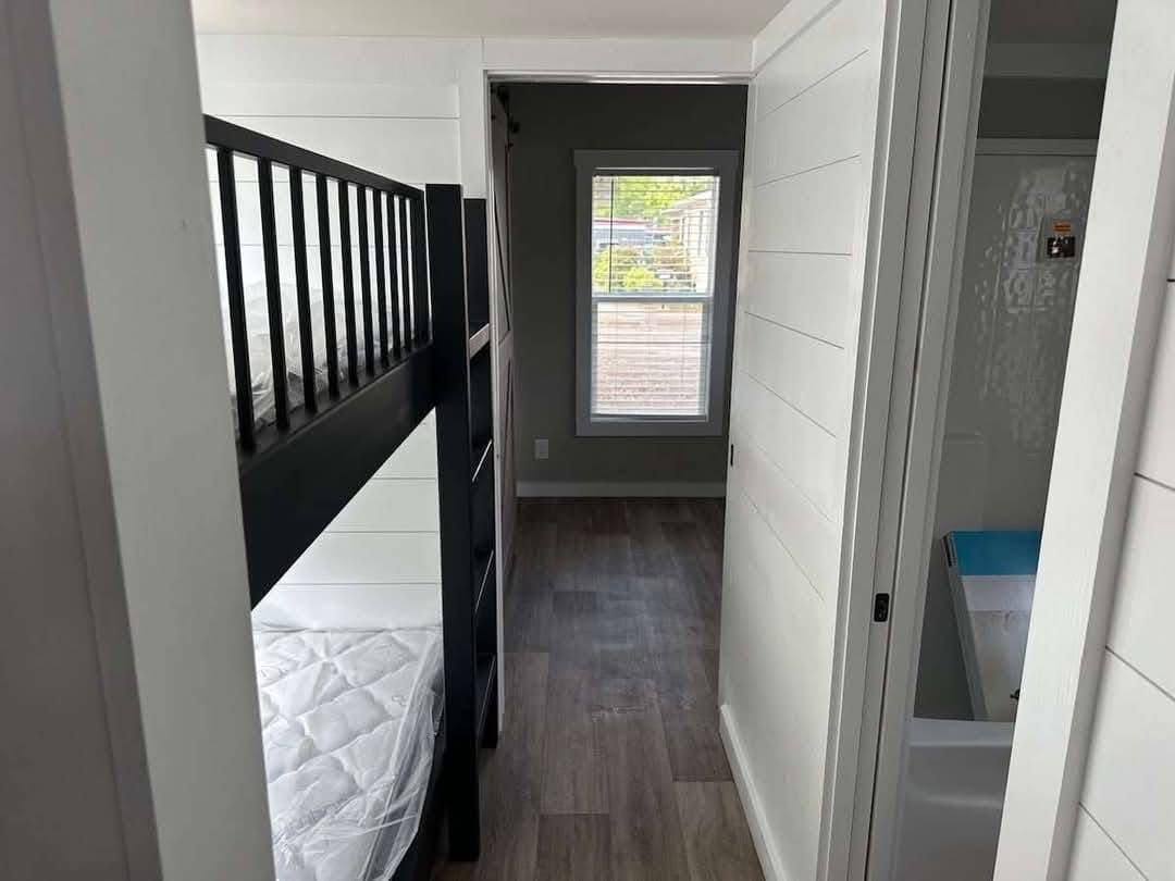 Narrow hallway with a dark wood bunk bed on the left, leading to a window with a view of greenery. A bathroom door is open on the right. Bright and tidy.