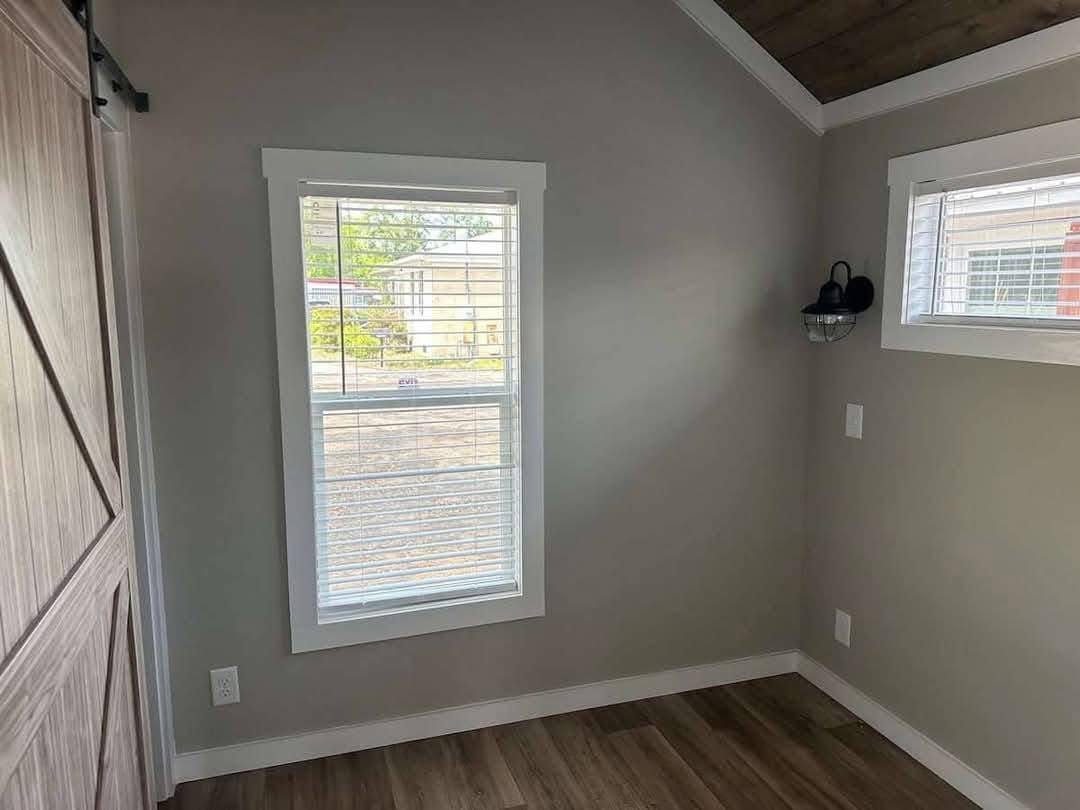 Small room with light gray walls and wood flooring. A large window with blinds on the left and a small window on the right, warm and inviting feel.