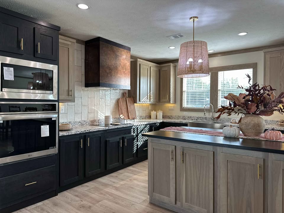 Modern kitchen with dark and light wood cabinets, granite countertops, and stainless steel appliances. A large pendant light hangs above an island with autumn-themed decor, conveying a warm, cozy atmosphere.