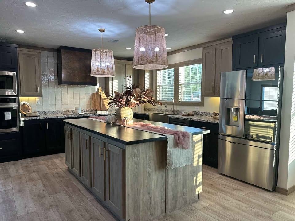 Modern kitchen with wooden floors, a central island, and a vase with dried flowers. Features stainless steel appliances, dark cabinets, and pendant lights, creating a warm and stylish ambiance.