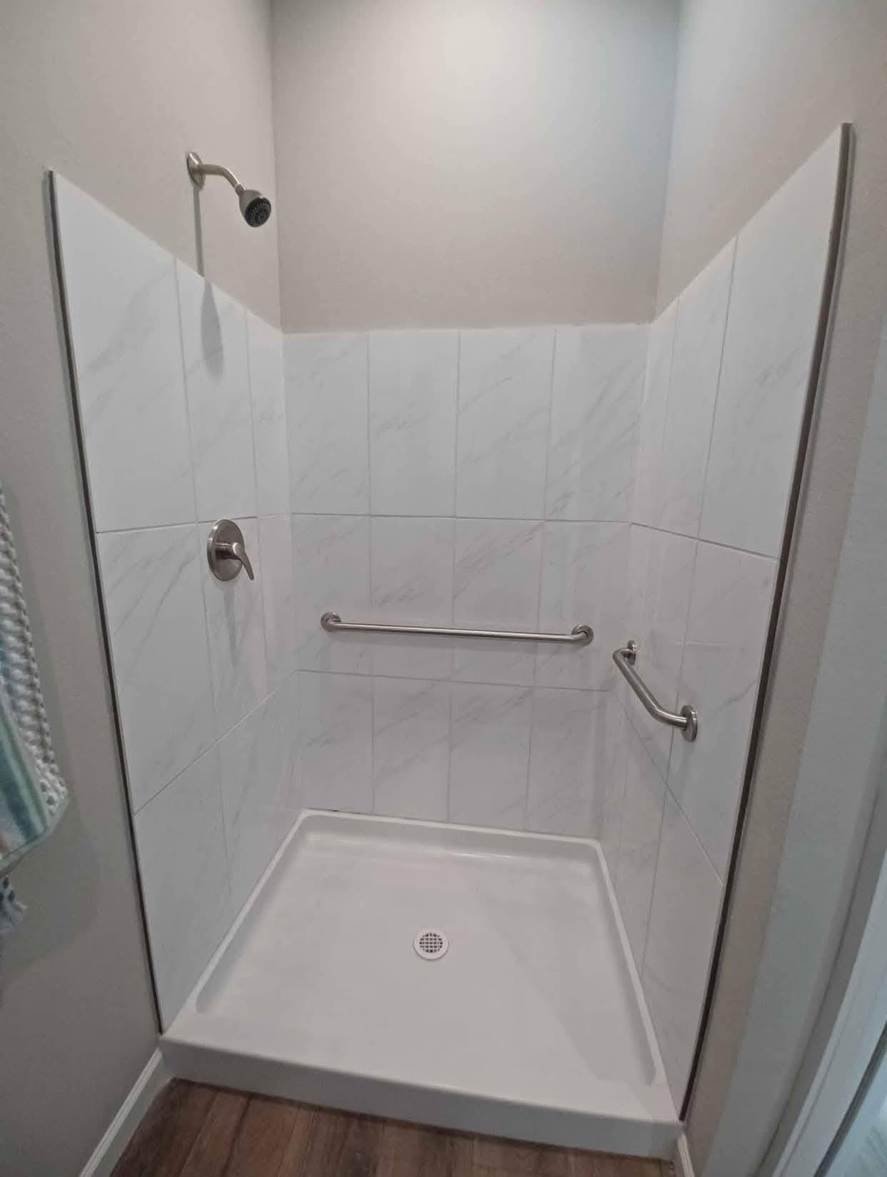 A small, accessible shower with white marble tiles, a detachable showerhead, and two metal grab bars. The floor is wooden, creating a clean, modern look.
