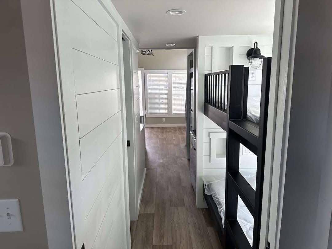 Narrow hallway with wooden floor, white walls, and a bunk bed on the right. Sunlight filters through blinds in the distant window, creating a cozy, minimalist feel.