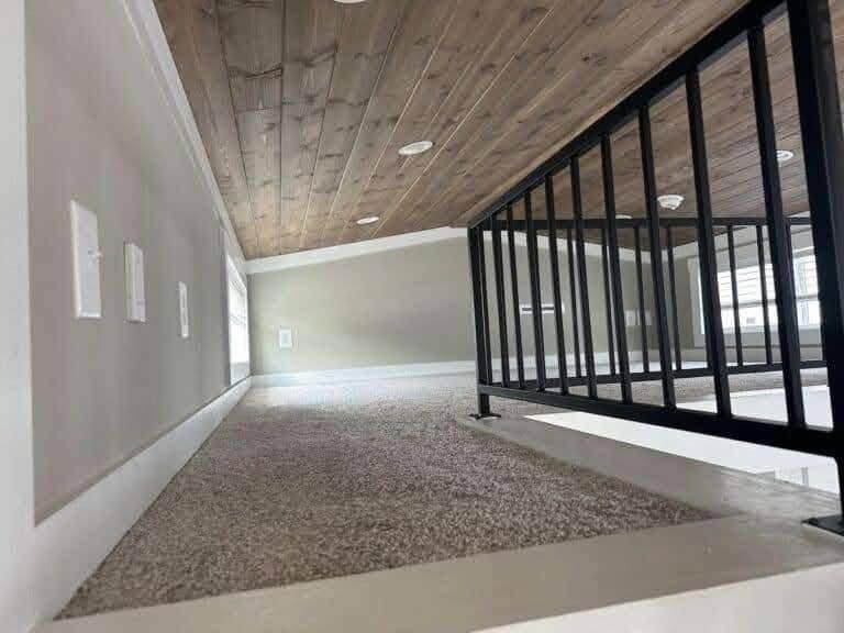 Low attic space with wooden ceiling and beige carpet floor. White wall plates are visible on the left, and a black railing leads to a windowed area. Cozy and compact.