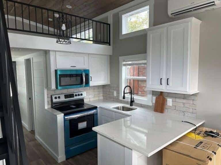 Modern kitchen with white cabinets, blue appliances, marble countertops, and tile backsplash. Upper loft with metal railing and warm, inviting atmosphere.