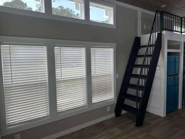 Interior of a modern tiny house with large windows and blinds, a wooden ladder leads to an upper loft, conveying a cozy and minimalist vibe.