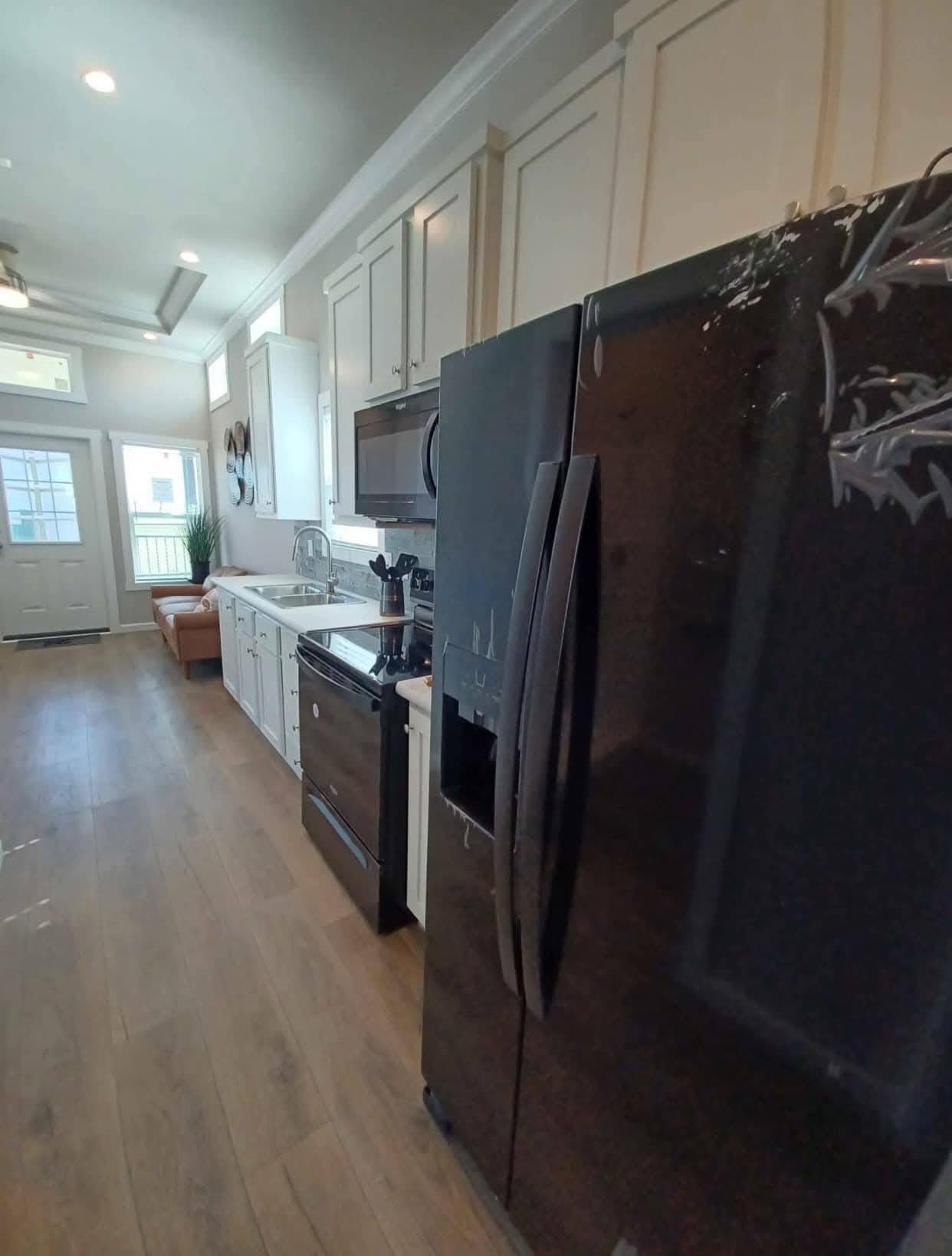 Narrow kitchen with wooden floors, white cabinets, and stainless steel appliances. Black fridge on the right. Bright and modern with a cozy sofa by a window.