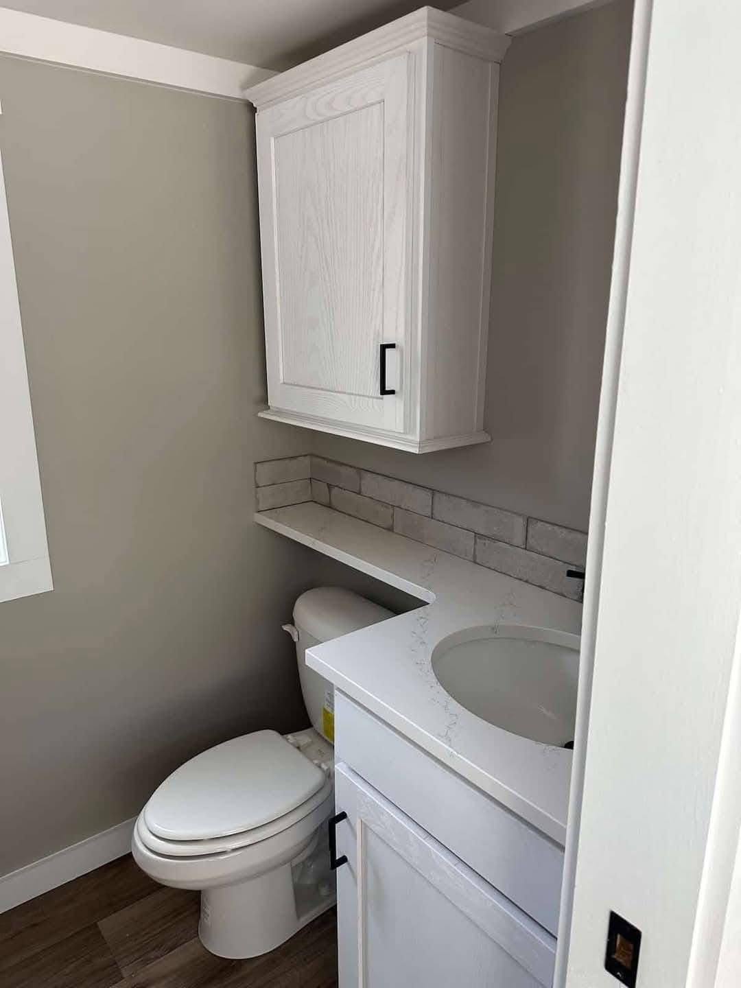 Compact bathroom with a white cabinet above a toilet, light gray walls, wood flooring, and a tiled backsplash along the sink counter.