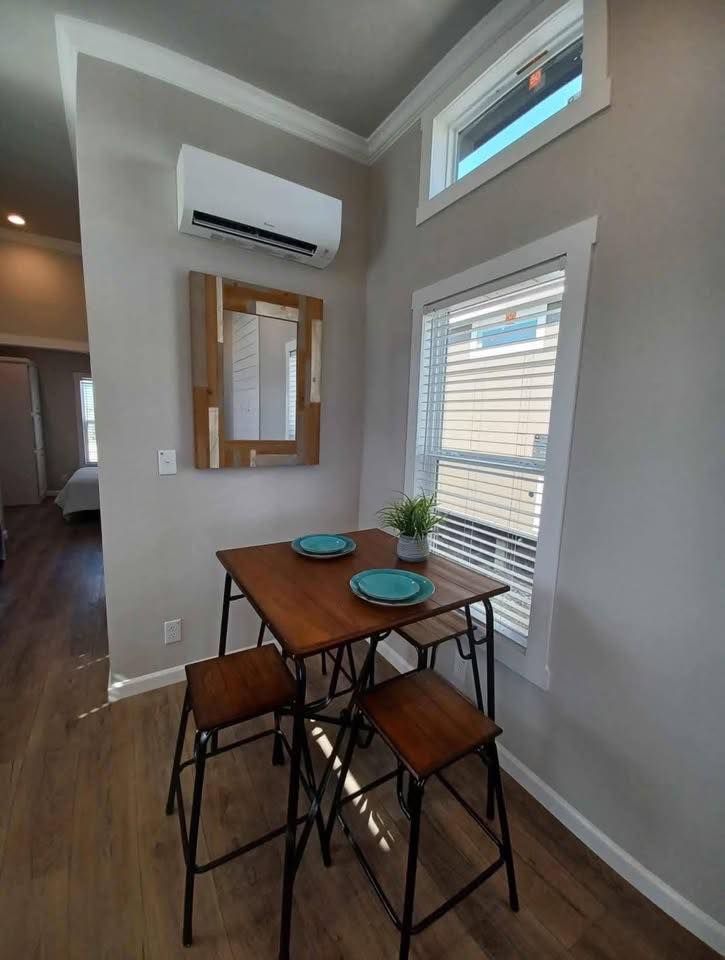 Compact dining area with a wooden table, two matching stools, and teal plates. A potted plant decorates the table. Large windows and a mirror enhance the space.