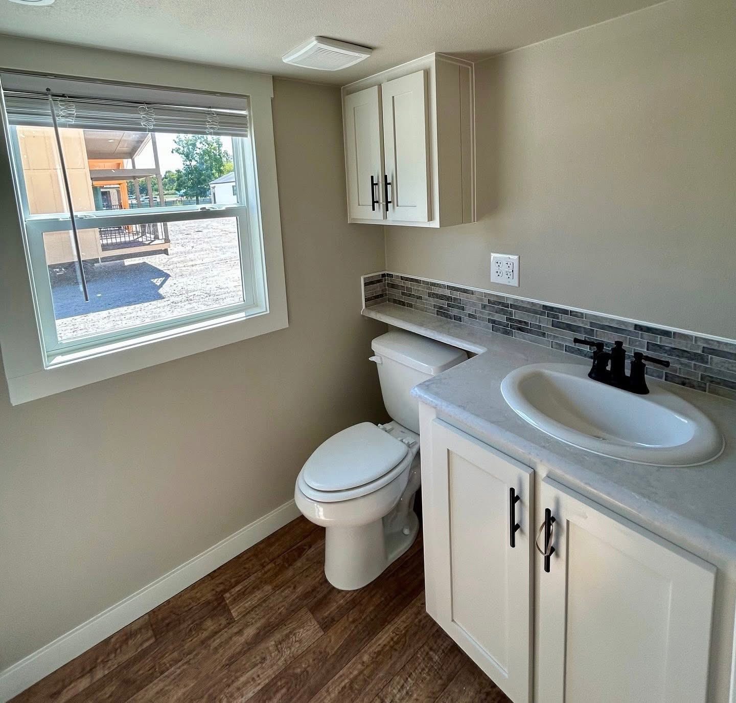 A small, bright bathroom with a window on the left, light wood flooring, a white toilet, and a sink vanity with black hardware and a mosaic backsplash.