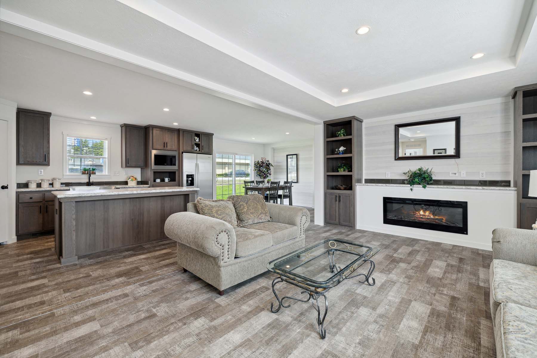 Spacious open-plan living room and kitchen. Features a cozy beige sofa, glass coffee table, modern fireplace, and wood cabinetry. Bright and inviting.