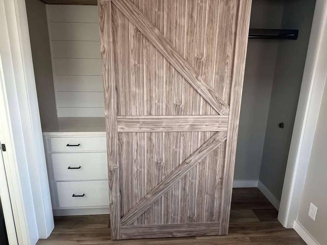 A wooden sliding barn door partially opens revealing a closet with white drawers and black handles on the left, conveying a rustic, modern vibe.