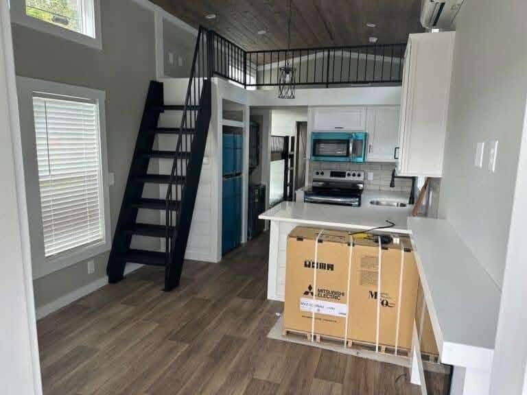 Cozy tiny house interior with wood flooring, featuring a sleek black metal staircase leading to a loft. Compact kitchen with white cabinets and blue appliances.