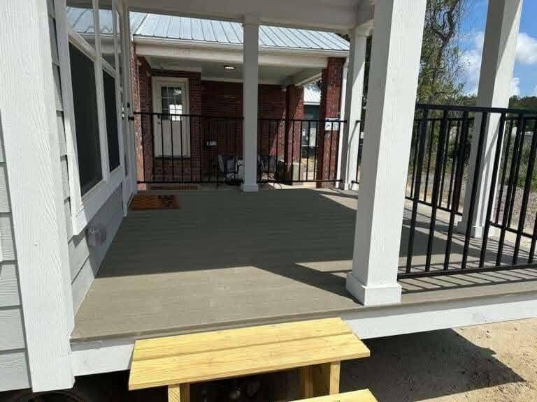 Covered porch with wooden steps leads to a cozy brick house. Black railing surrounds the porch, and a welcome mat is placed near the door. Sunny day.