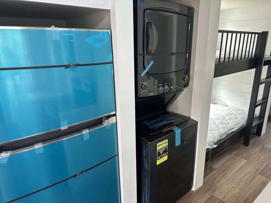 Stacked black washer and dryer beside a large blue refrigerator. Adjacent, a black bunk bed with white mattress is on a wooden floor.