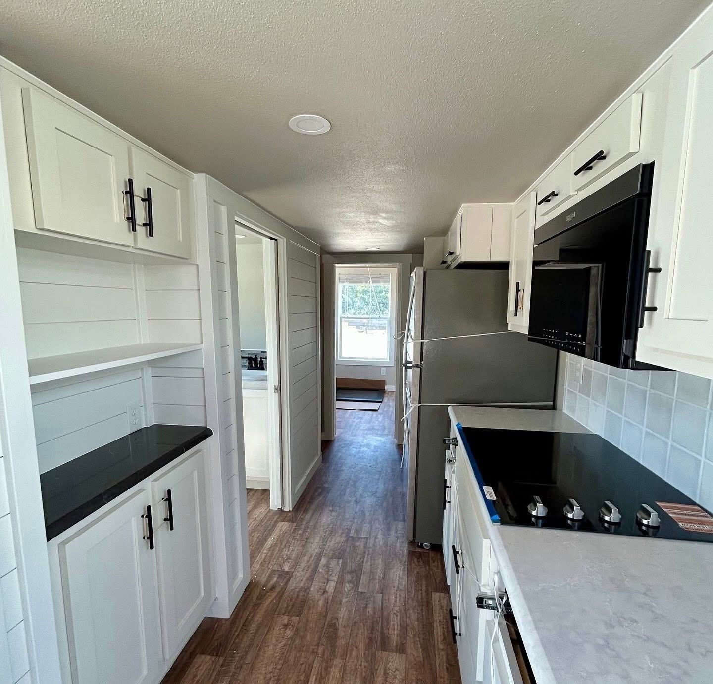 Compact kitchen with white cabinets, black handles, and stainless steel appliances. Wood flooring adds warmth. A window in the back lets in natural light.
