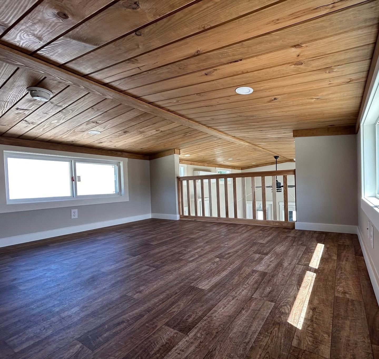 Cozy loft with warm wooden flooring and ceiling, featuring a railing overlooking a bright space. Two windows bring in natural light, creating a serene atmosphere.