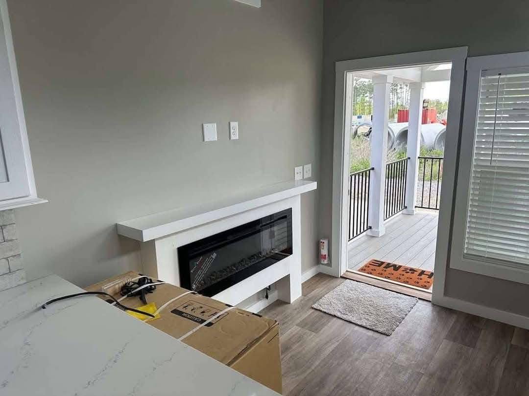 Modern room with light gray walls, laminate flooring, and a wall-mounted fireplace. A sliding door leads to a porch with black railings and a welcome mat.