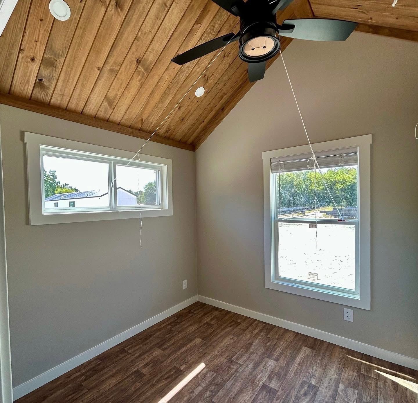 Small room with light brown walls and a wooden ceiling featuring a black fan. Two windows illuminate the space, highlighting the wood-patterned floor. Cozy and airy atmosphere.