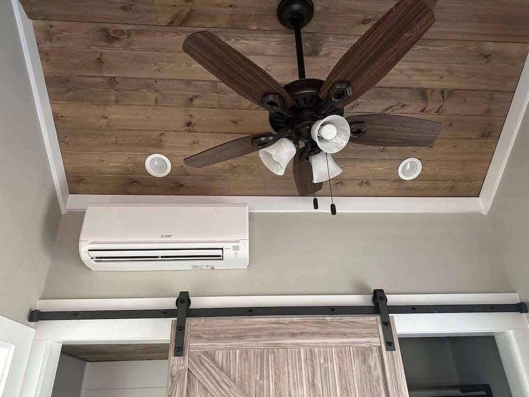 Wooden ceiling with a dark wood fan, white light fixtures, and a recessed light. Below is a sliding barn door and a sleek wall-mounted AC unit. Rustic and modern.