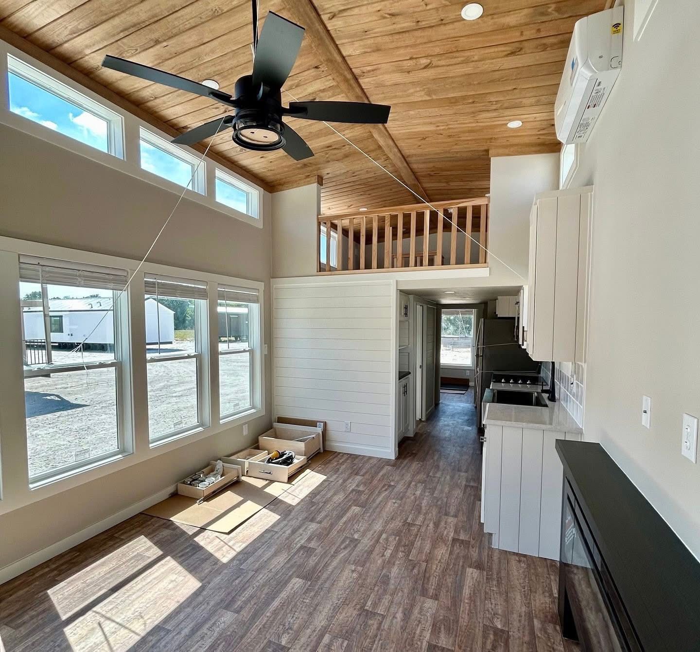 Bright tiny home interior with wooden ceiling and floor, large windows, and a black ceiling fan. Loft railing overlooks the compact kitchen below.